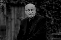 A black-and-white photograph of Salman Rushdie sitting on a park bench.