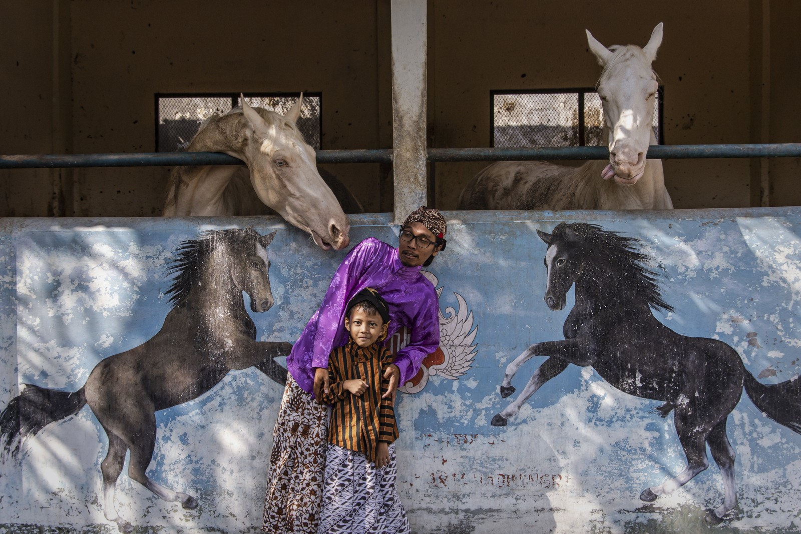 A man and child pose along a wall near horses. The man leans away as a horse leans toward him. 