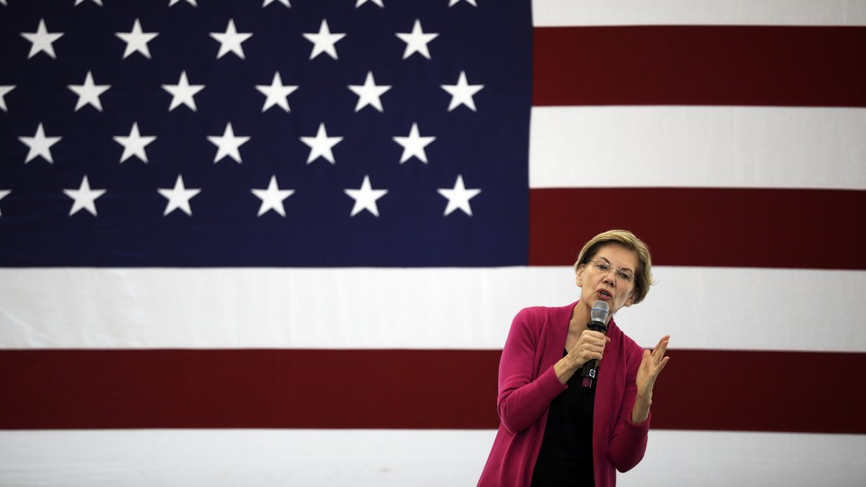 Elizabeth Warren, wearing a pink cardigan, speaks into a microphone onstage at an event. A giant American flag is displayed behind her.