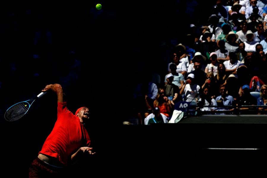 A tennis player takes a large swing at a tennis ball, as a crowd watches from behind.