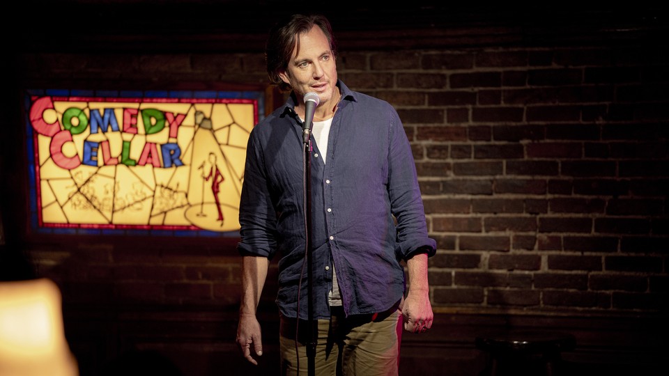 A man in a blue button down and khakis stands in front of a microphone with a brick wall and a sign reading 'Comedy Cellar' behind him