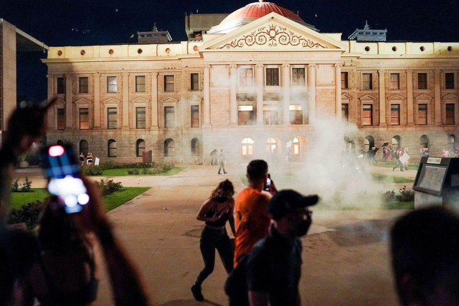 Several people are scattered by a cloud of tear gas in a plaza outside a capitol building.