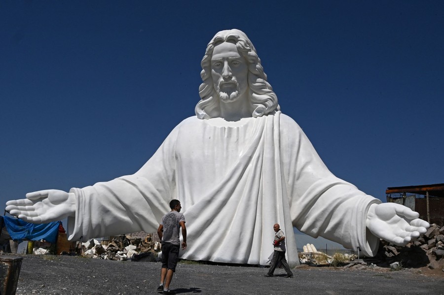 A couple of people walk around the base of the upper half of a very tall, white sculpture of Jesus Christ.