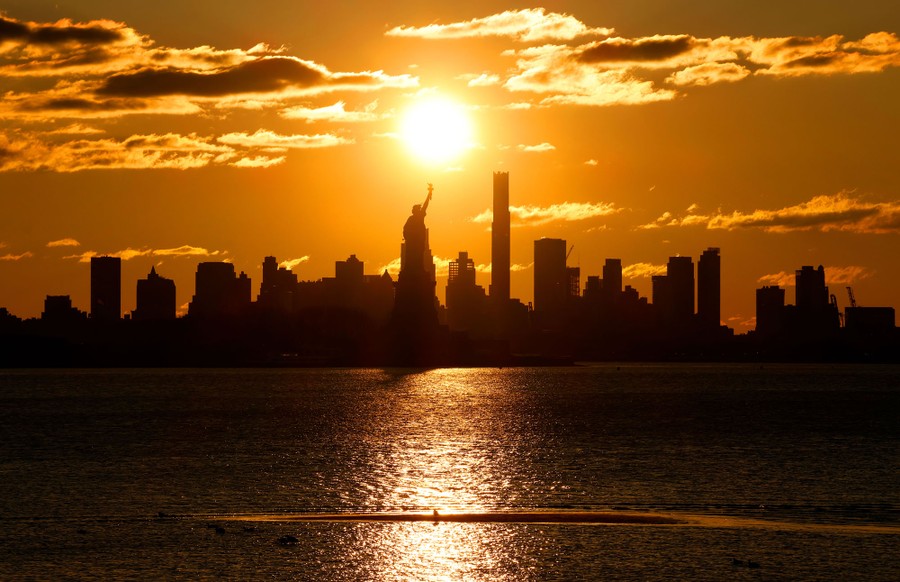 The sun rises behind the Statue of Liberty and the skyline of New York City.