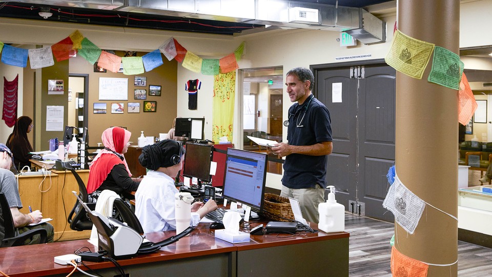 A man with a stethoscope around his neck stands in front of people sitting at computers in a doctor's office.