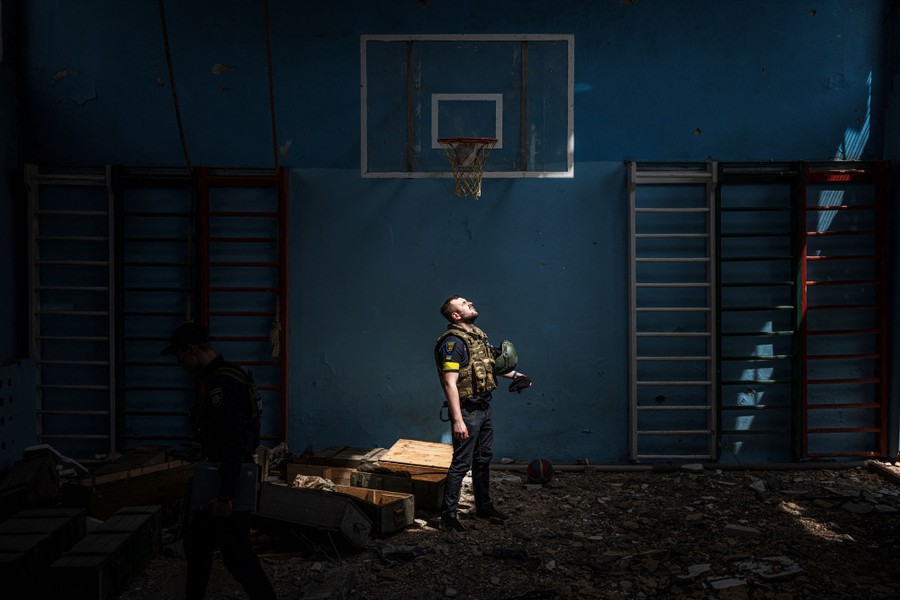 A police officer stands inside a dark, damaged school gymnasium, looking up toward sunlight pouring in through a hole in the roof.