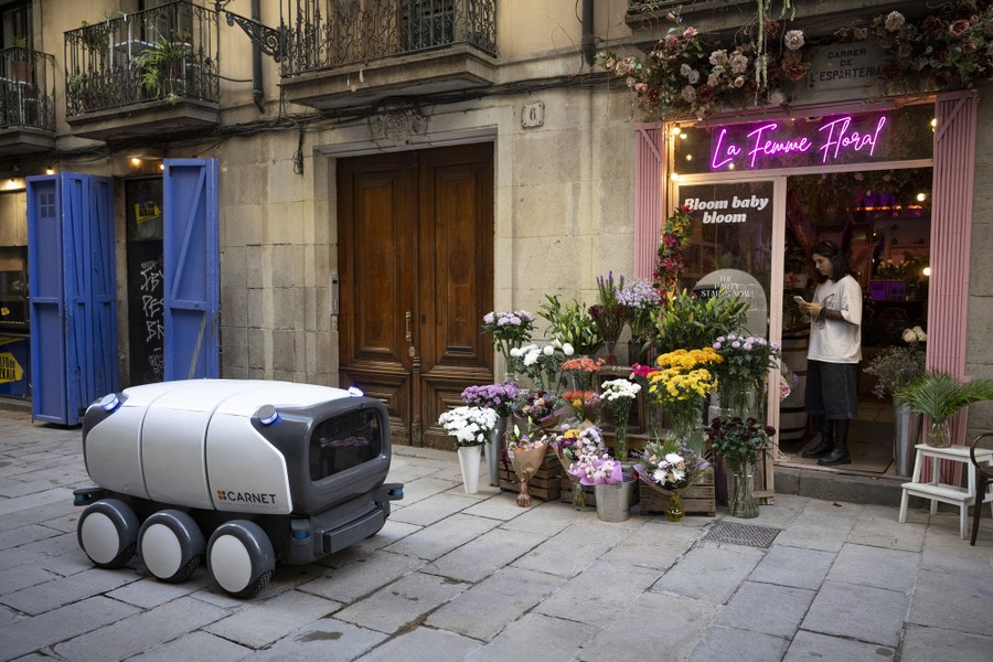 A small six-wheeled autonomous vehicle rolls past a flower shop.