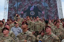 A group of soldiers stands in front of a Jumbotron showing Donald Trump at a podium above them