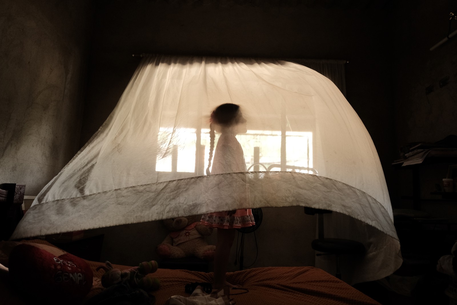 A young girl stands on a bed, beneath a billowing curtain.
