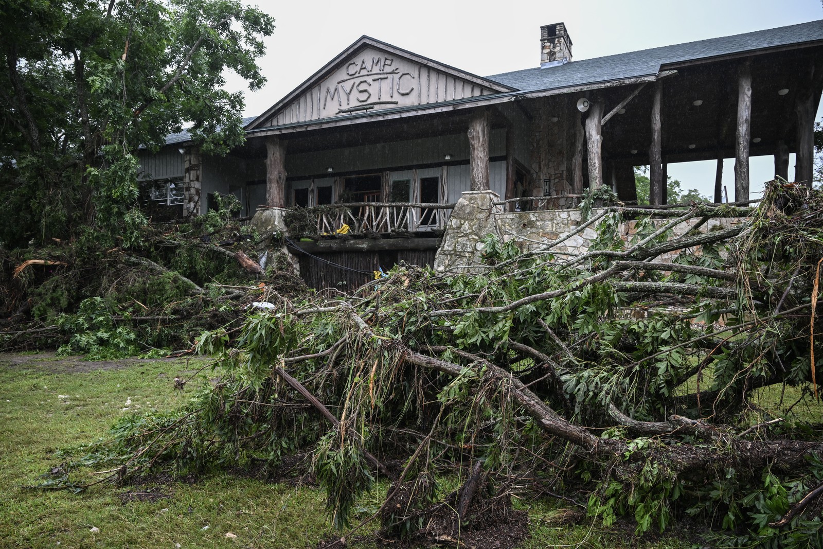 Broken trees and branches lie scattered in front of a large camp building that has a sign reading 