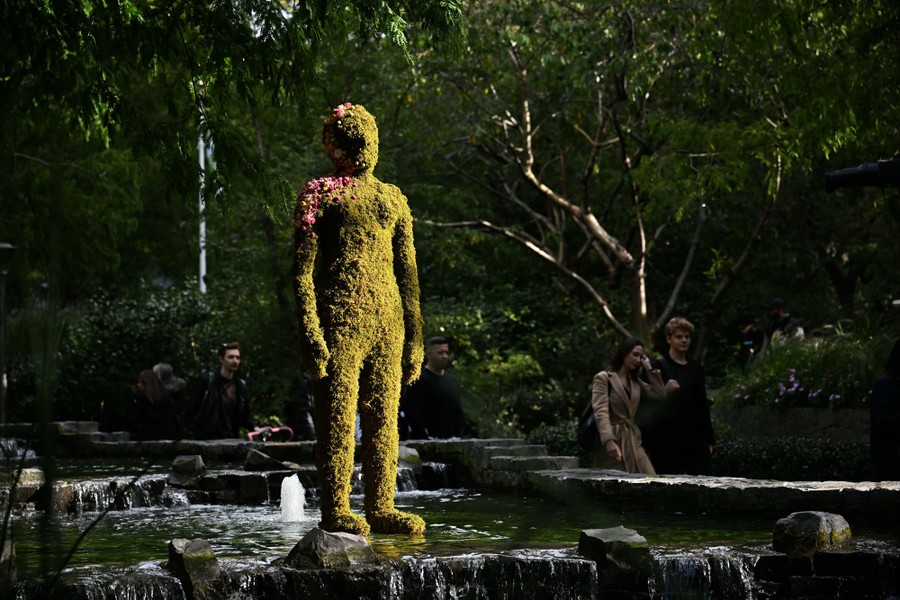A sculpture stands in a pool of water, the same shape and size as a human, covered in moss and flowers.