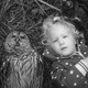 A black-and-white photo of an owl and a child lying in the grass, staring into the lens of the camera.