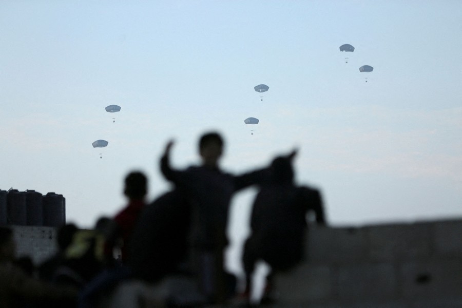 Several people are seen in the foreground, looking toward six parachutes in the sky beyond.
