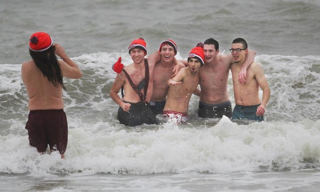 Six men in swimming trunks stand in the ocean. One of them has his back turned and is taking a picture of the other five. Some of them are wearing winter hats and mittens.