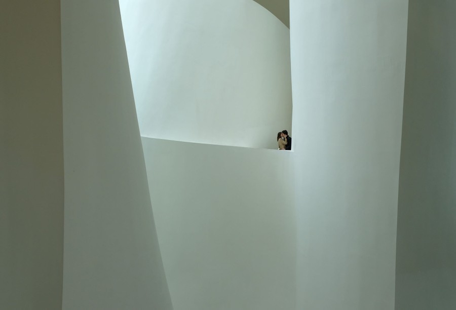A couple share a kiss standing along a balcony inside an atrium in a modern museum.