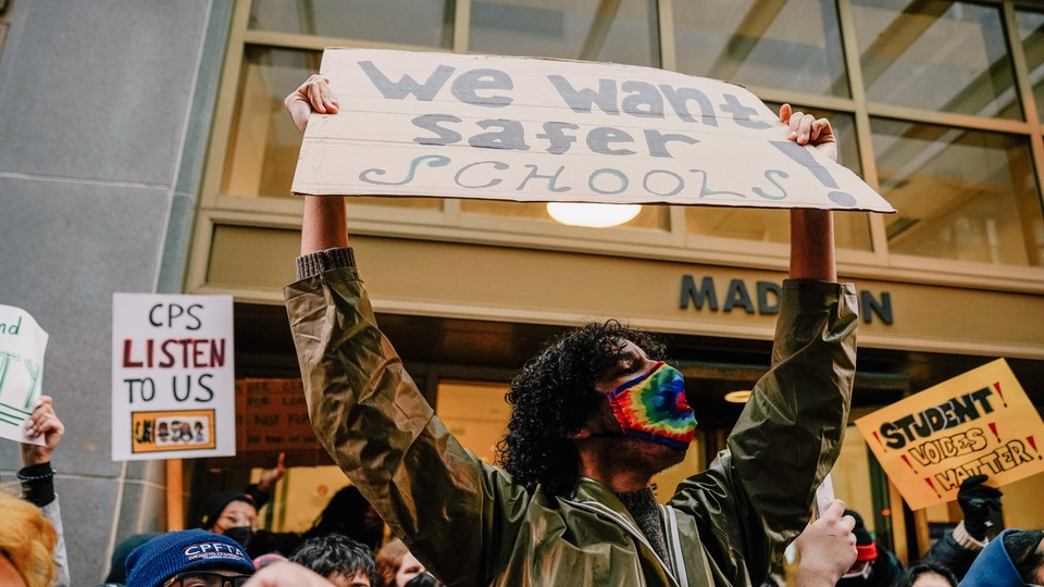 A young man wearing a rainbow mask holds up a sign that says "We want safer schools." In the crowd behind him, other signs read: "CPS Listen to Us" and "Student Voices Matter"
