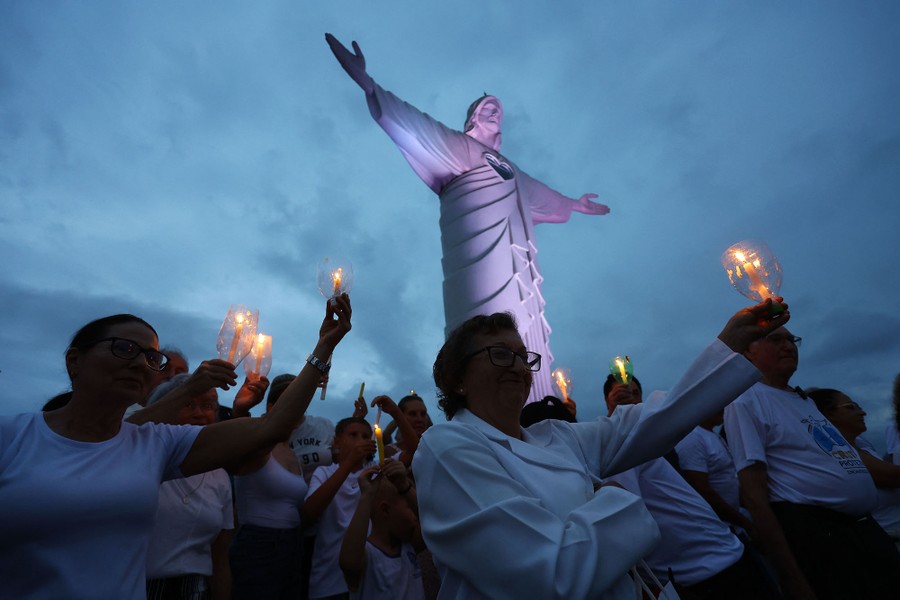 A group of people hold up candles during a prayer beneath a large statue of Jesus Christ.