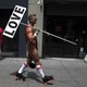 A man in roller skates and an over-the-shoulder thong holding a sign that reads "LOVE"