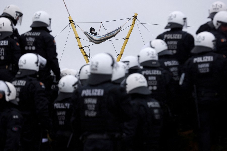 A person sits in a hammock suspended high above the ground from ropes and logs as a large group of riot police gather below.