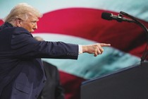 Color photo of Trump pointing with his mouth open, beside a miced-up podium and a large American-flag background.