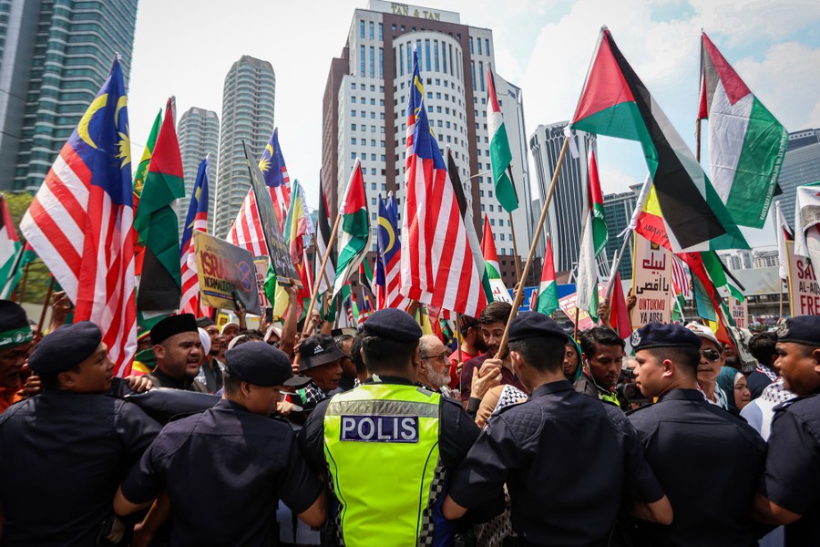A line of police officers stands in front of a crowd of protesters waving flags of Malaysia and Palestine.