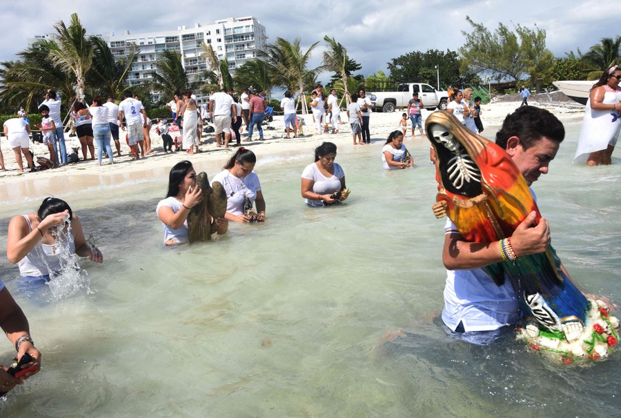 People wade in the surf on a beach, some carrying religious figures.