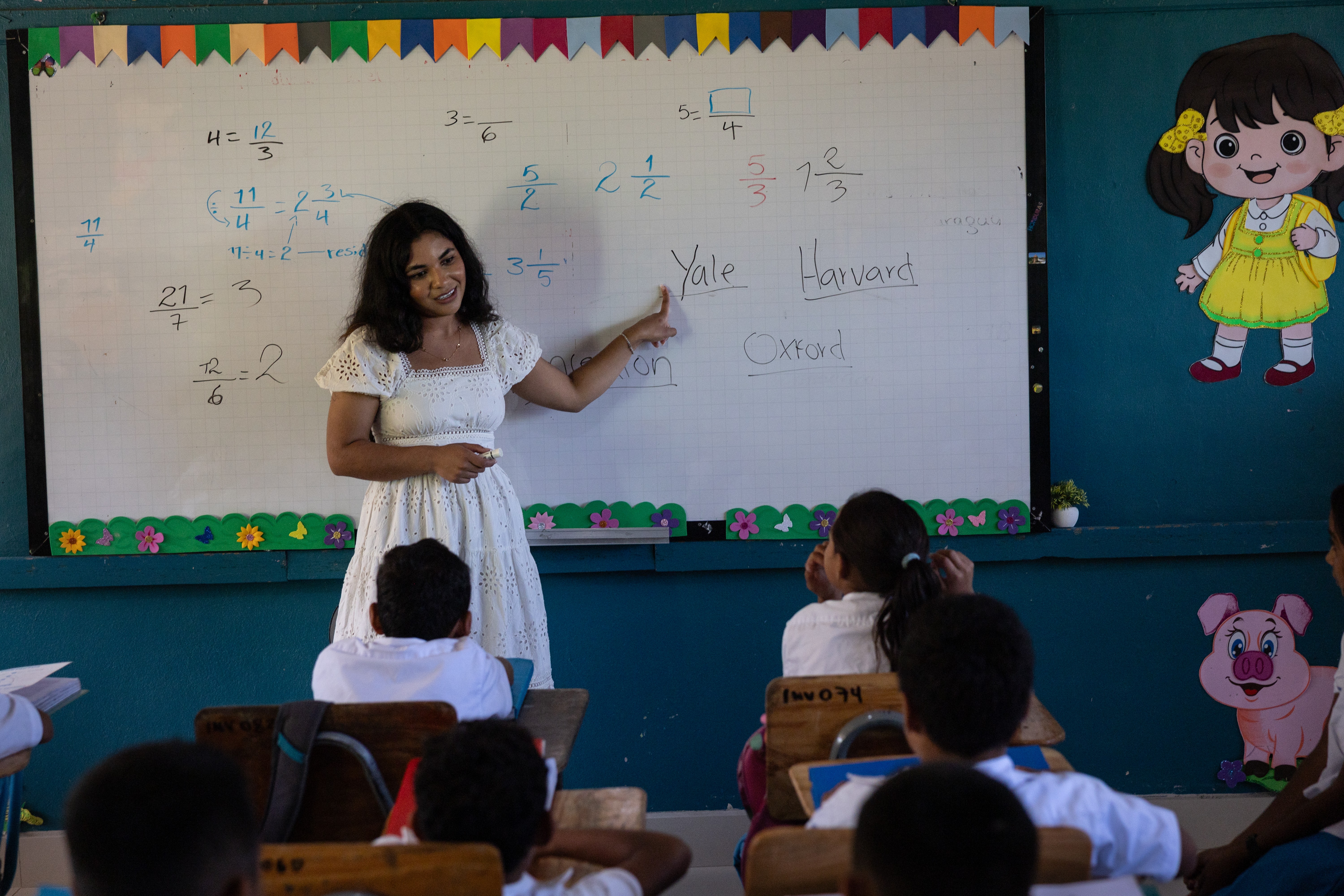 A young woman stands in front of a whiteboard, speaking to students in a classroom.