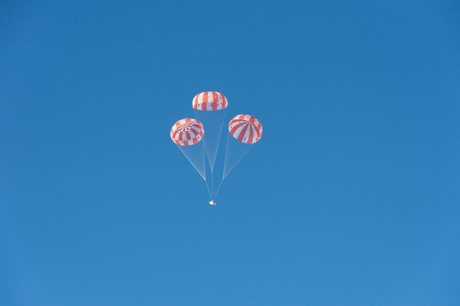 A small space capsule dangles from three large parachutes in a blue sky.
