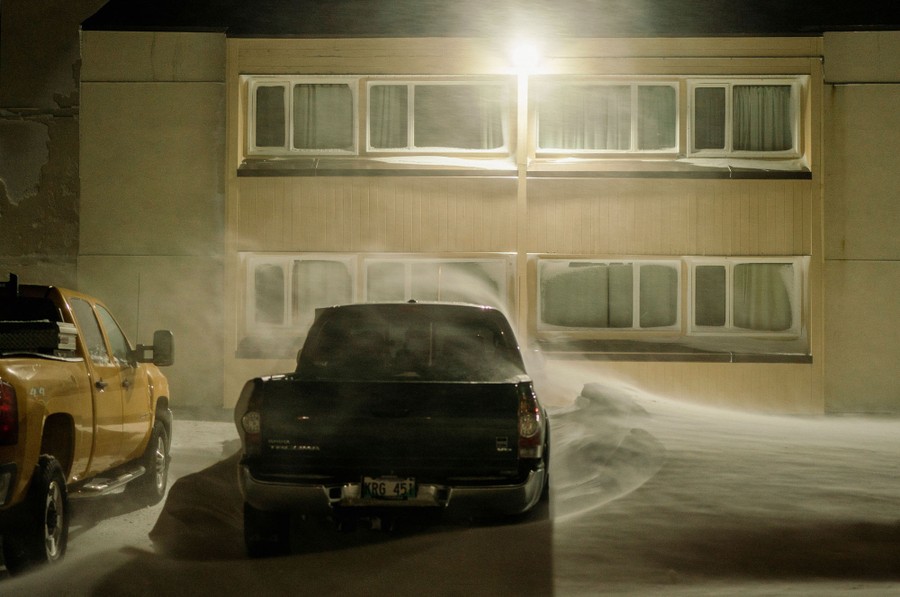Snow drifts around a parked pickup truck outside a building.