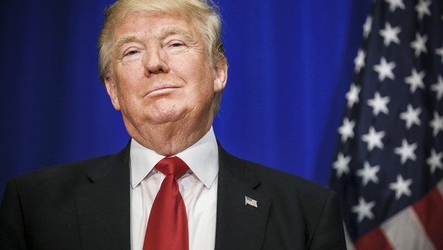 Trump in suit smirks beside an American flag
