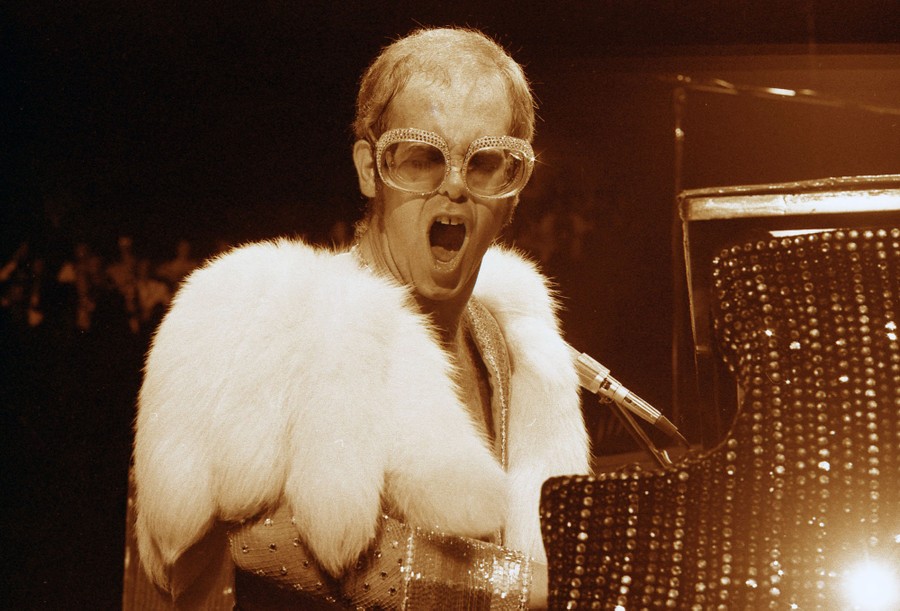 The performer Elton John, playing a piano and singing while wearing fur over his shoulders and large sparkly glasses