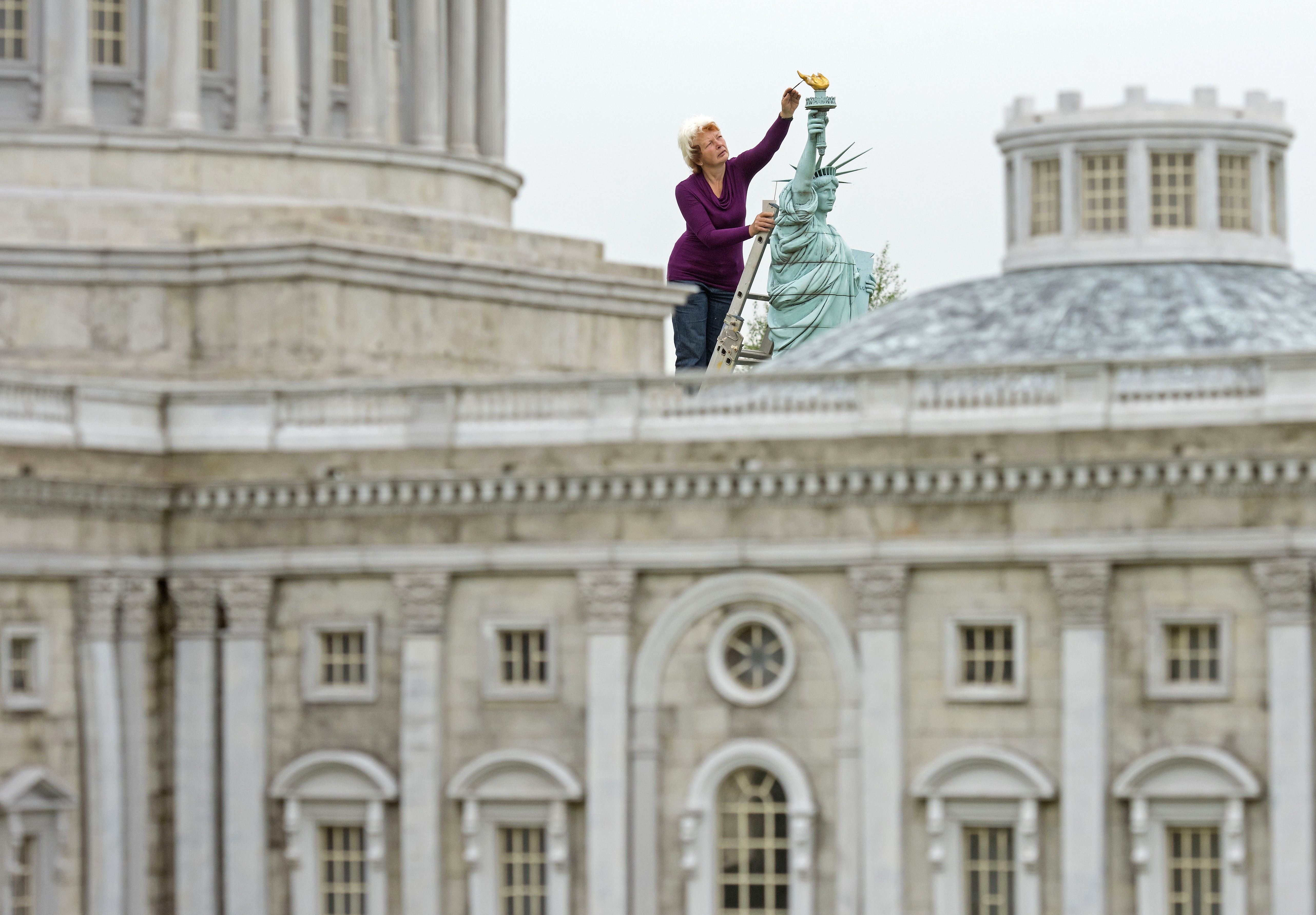 A person stands on a ladder, working on a small-scale replica of the Statue of Liberty.