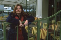 A woman in a blue coat and red scarf with red hair leans on a green railing in an empty outdoor restaurant patio.