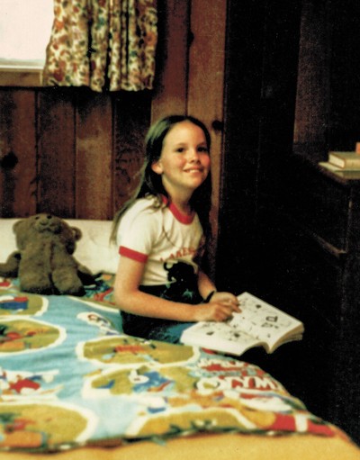 color photo of smiling child holding book and sitting on bed next to stuffed animal