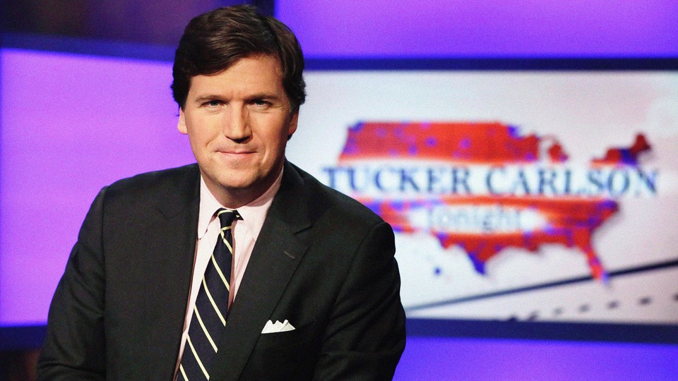 A photograph of Tucker Carlson, wearing a suit and tie, and smiling into the camera in front of his former show's logo
