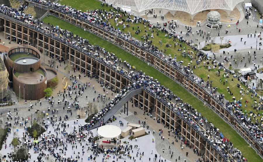 An aerial view of crowds of people walking through a fairground, and on top of a tall ring structure