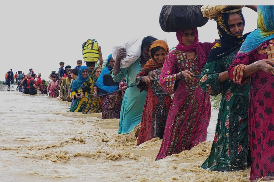 A line of women and men carry bundles through fast-flowing knee-deep floodwater while holding a rope.