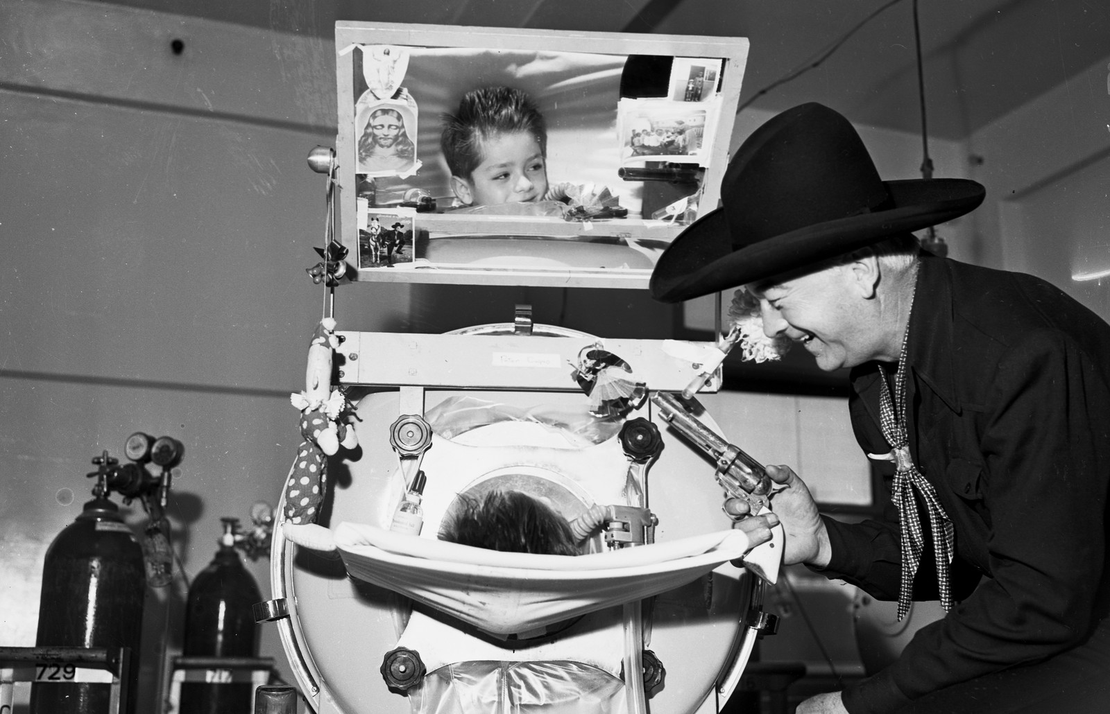 A performer dressed as a cowboy leans over qand smiles at a child who is resting inside an iron lung.