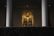 A color photograph of a White House door at night, seen behind a hedge and between two columns.