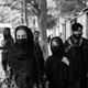 Black-and-white photo of two women with covered faces in a street. Several men are in the background behind them.