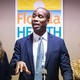 Florida Surgeon General Joseph Ladapo delivers a speech in a blue suit as two women watch behind him.