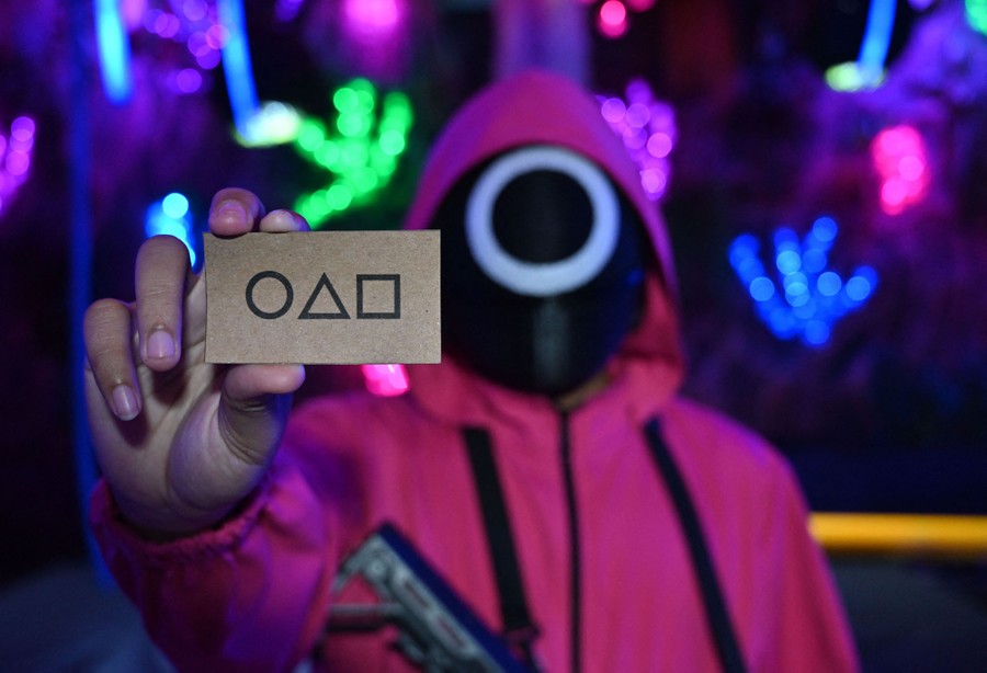 A person dressed in an outfit from the Netflix series "Squid Game" holds a card aloft. The card has a circle, a triangle, and a square printed on it.