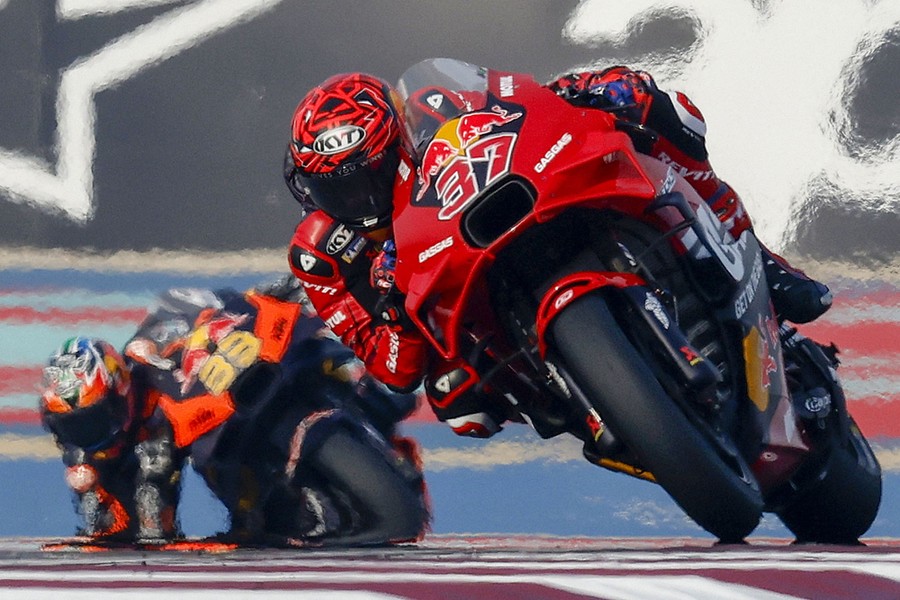 Two MotoGP motorcycle racers lean into a turn on a racetrack, with a heat shimmer distorting the background.
