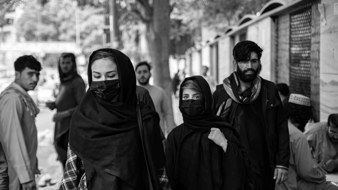 Black-and-white photo of two women with covered faces in a street. Several men are in the background behind them.