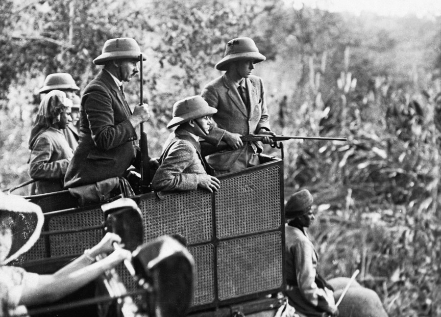 Several men wearing pith helmets and carrying long guns ride in a basket atop an elephant.