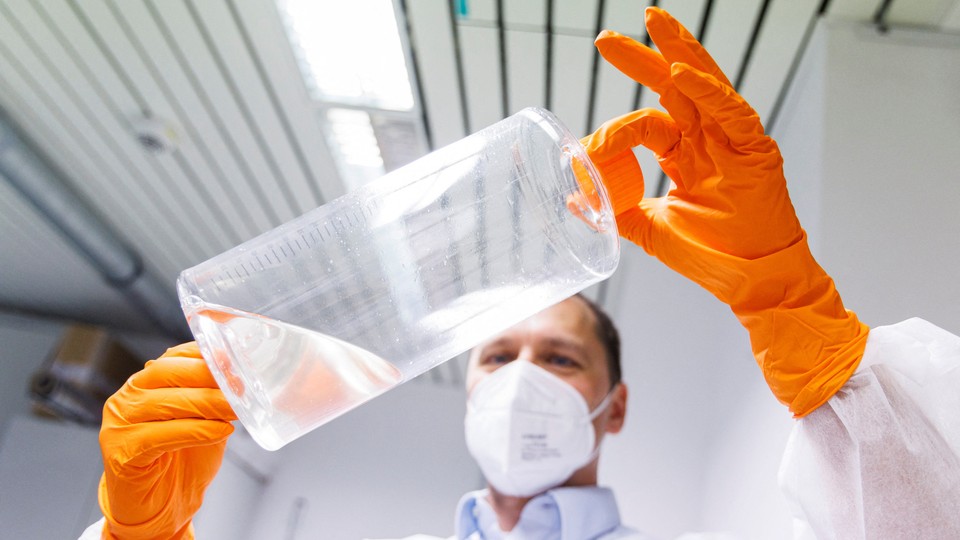 An employee of the German company that makes the Jynneos vaccine holds up a clear bottle with some liquid in it. The employee is wearing a medical face mask and orange gloves.