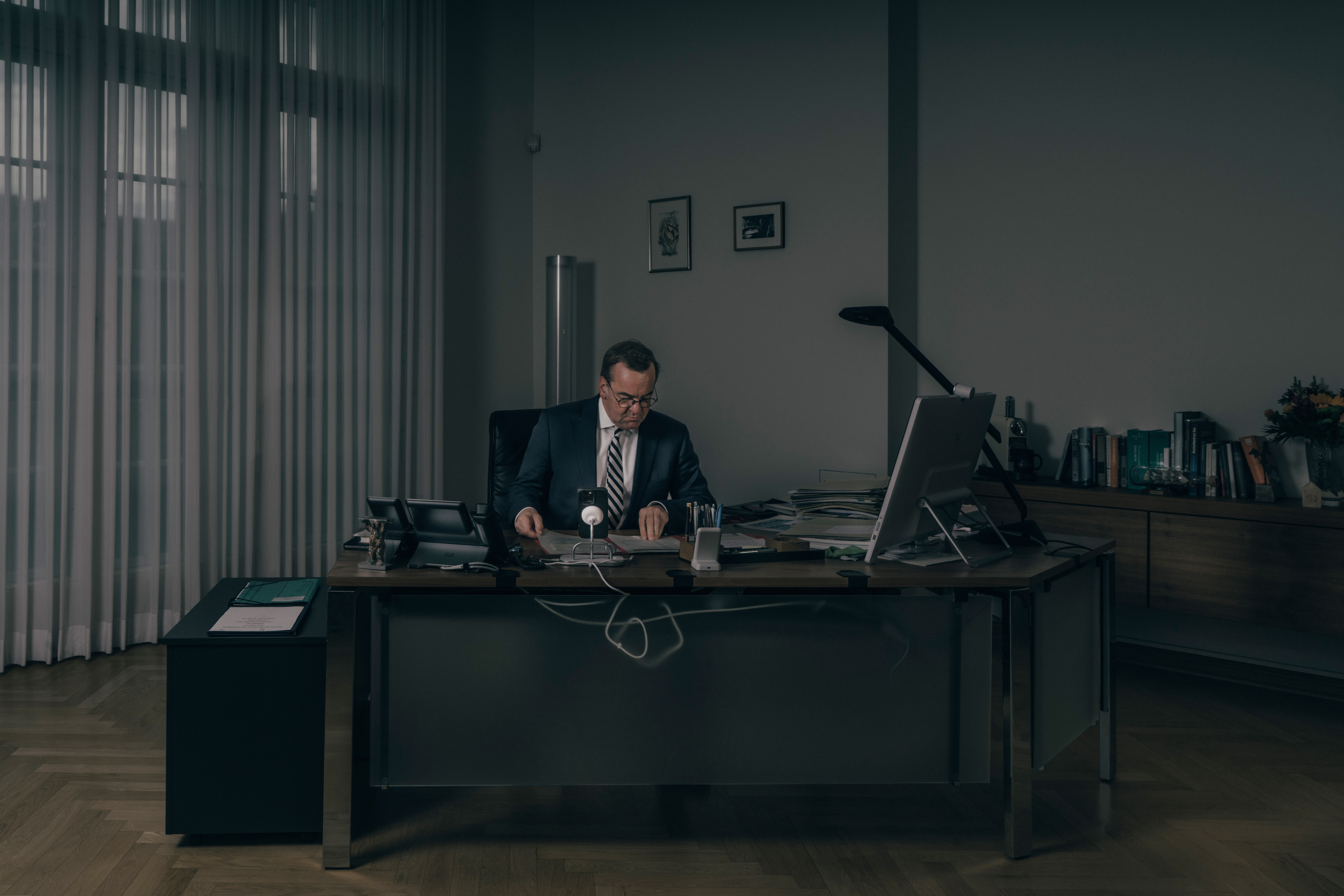 photo of man wearing suit and working at desk in large office with floor-to-ceiling curtained window behind