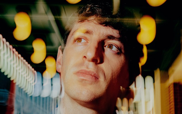 A photo of Alex Edelman surrounded by blurred lights outside of Hudson Theatre in New York