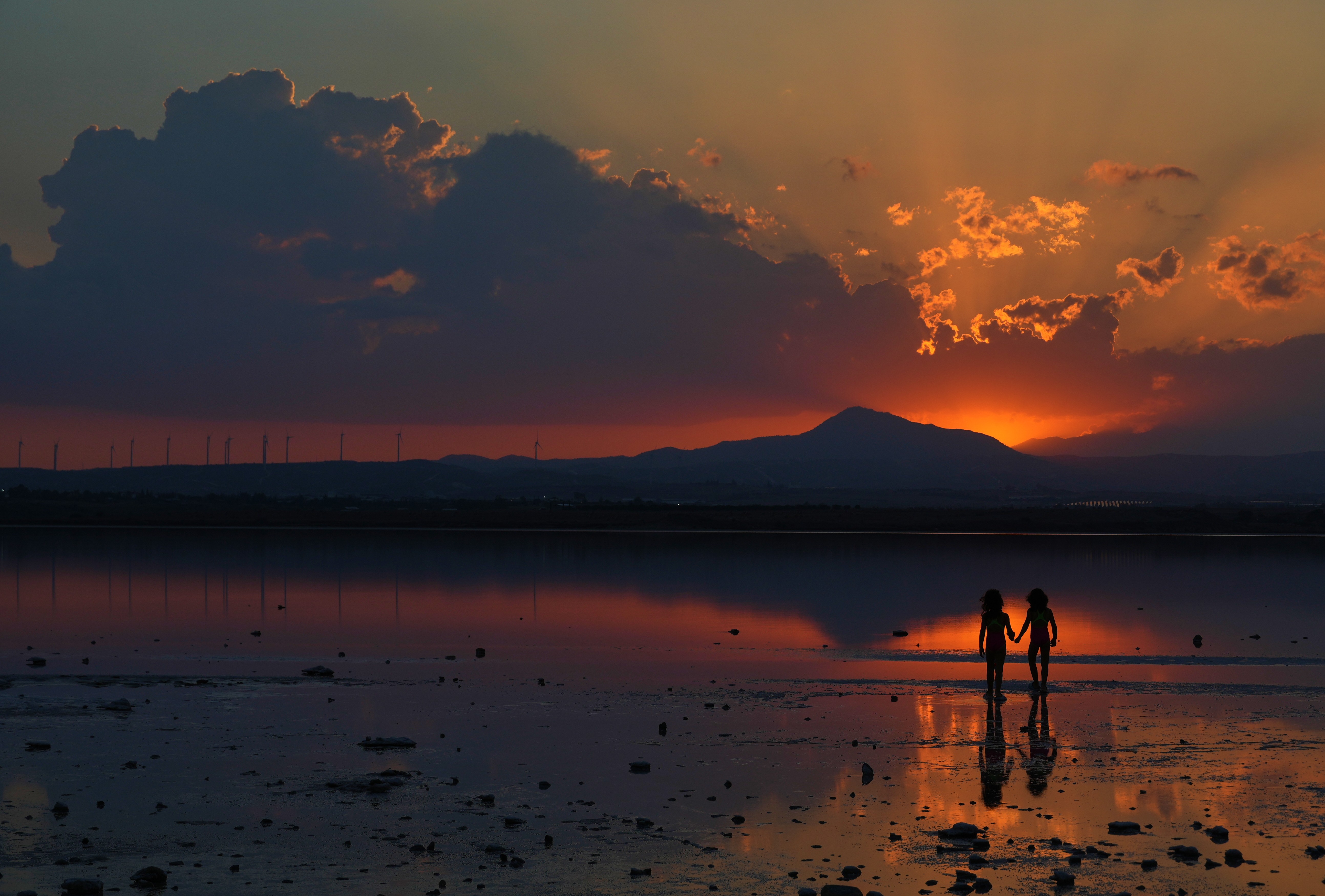 Two girls stand near the shore of a salt lake, seen in silhouette at sunset.