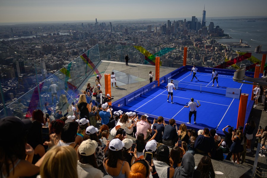 People play pickleball in front of an audience on a small court set up on a high observation deck above New York City.